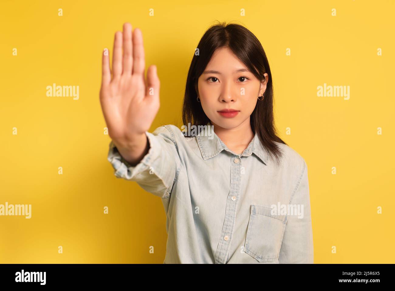 Young Asian lady gesturing STOP, showing palm at camera on yellow studio background Stock Photo