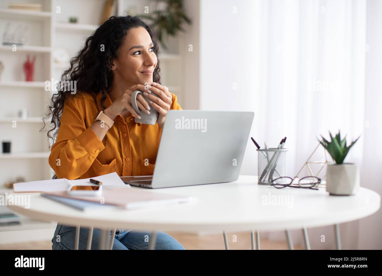 Dreamy Businesswoman Using Laptop And Holding Coffee Cup Indoors Stock Photo
