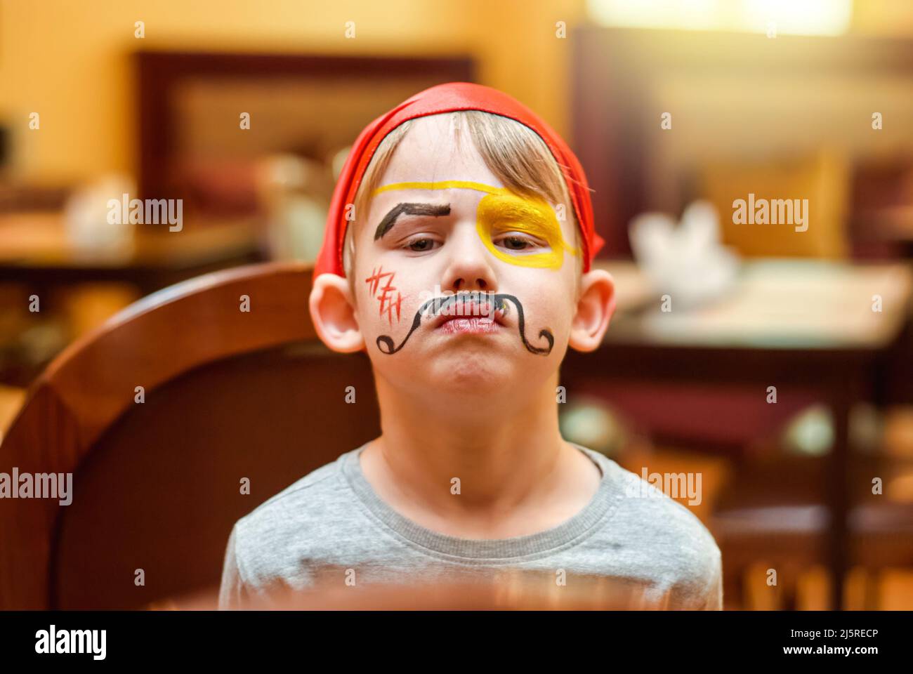 Preschooler feels unhappy after animator painting on face made at children party. Portrait of blond boy with fake mustache on blurred background close Stock Photo