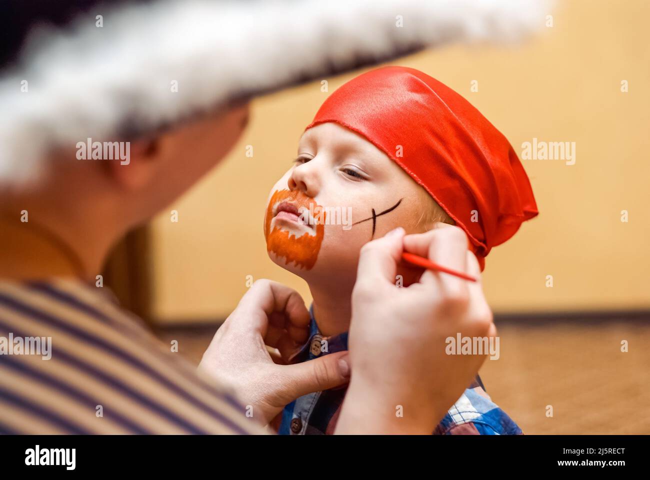 Animator makes pirate painting on face of toddler at children birthday party. Cute boy in red bandanna prepares for celebration close view Stock Photo