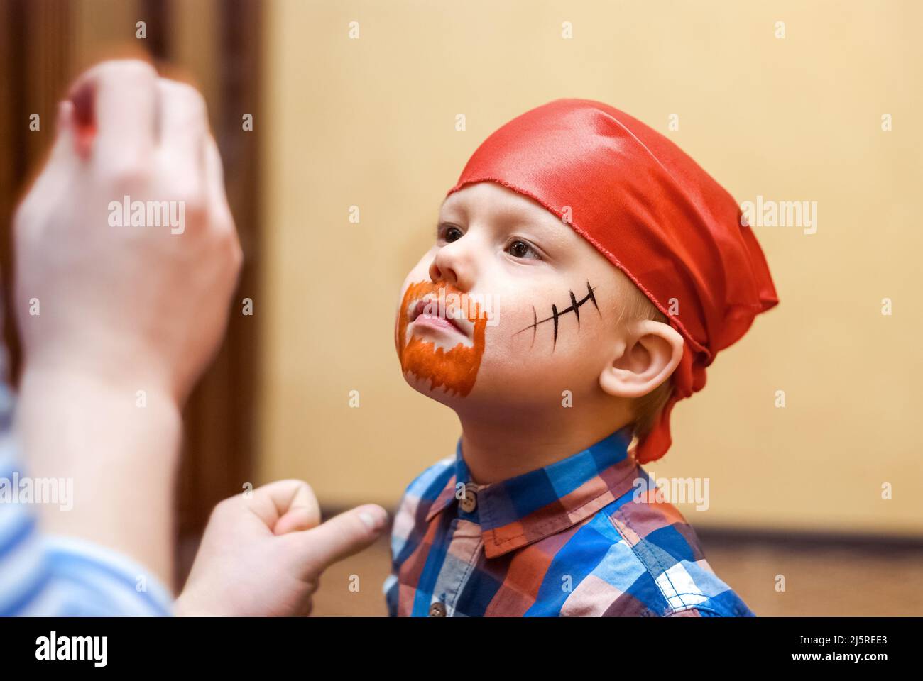 Animator makes pirate painting on face of toddler at children birthday party. Cute boy in red bandanna prepares for celebration close view Stock Photo