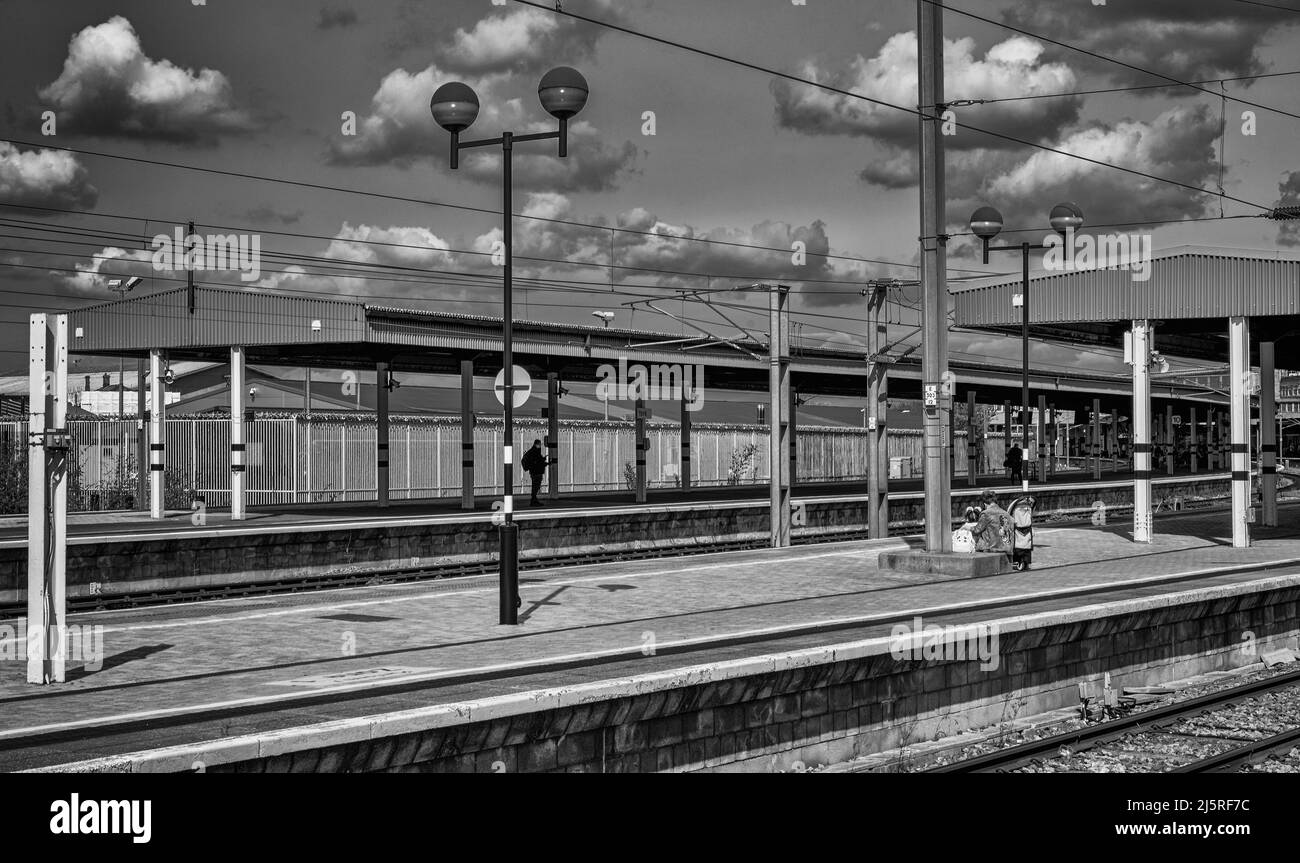 A woman holding a child sits on a railway station platform. A cloudy sky is above. Stock Photo