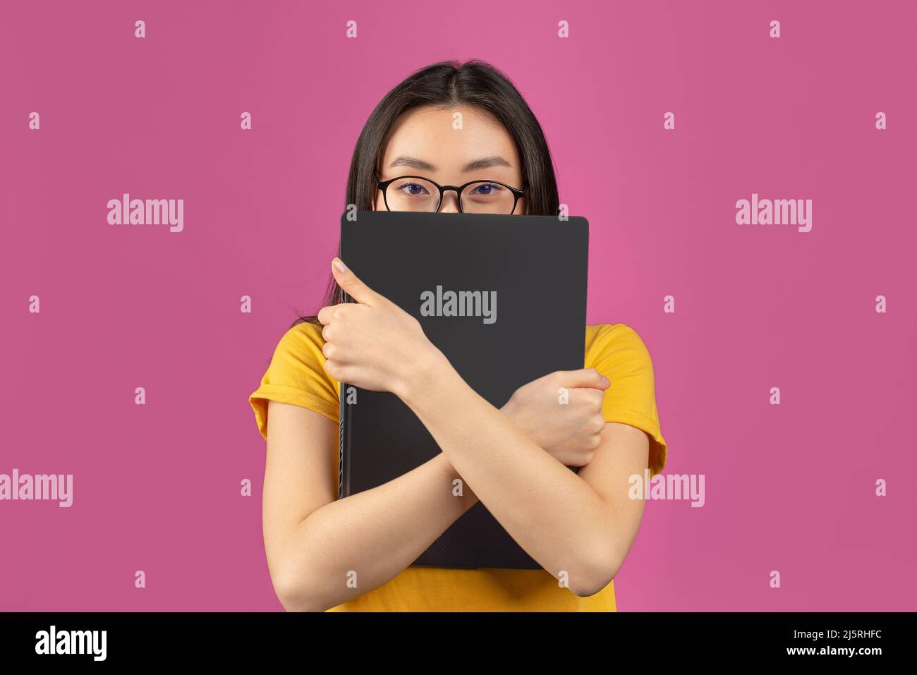 Asian female student in glasses hiding behind laptop, studying for college exams online, standing on violet background Stock Photo