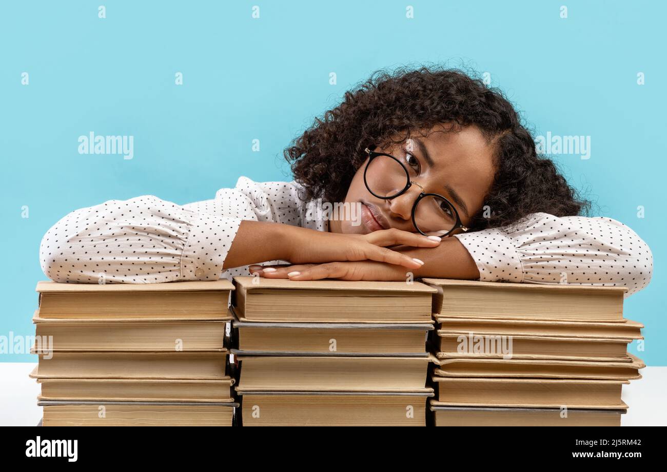 Sleepy young black female student leaning on big stacks of books, tired of getting ready for exam, feeling bored Stock Photo