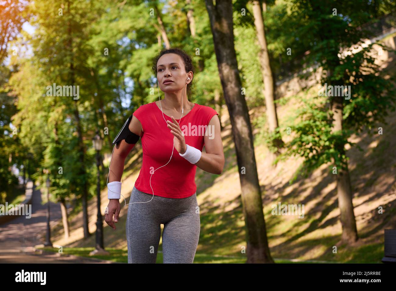 Attractive strength muscular multi ethnic woman with healthy beautiful aesthetic fit body in tight sportswear with earphones and smartphone holder run Stock Photo