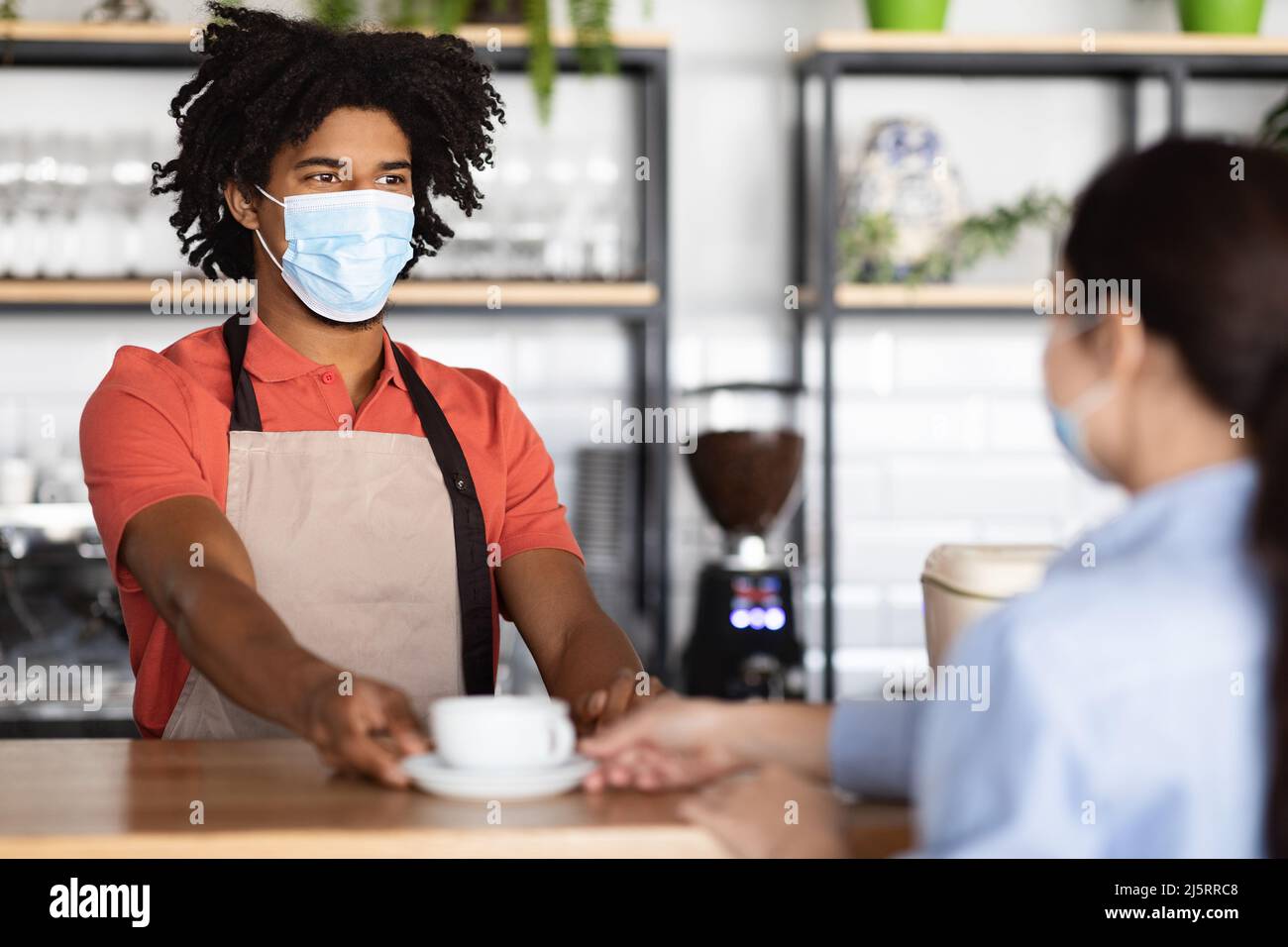 Smiling young african american curly male waiter in protective mask and apron giving fresh coffee to client Stock Photo