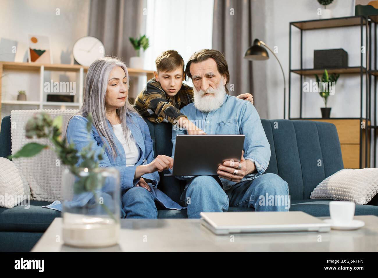 Pleasant happy smiling family of grandparents and grandson sitting on sofa at home and making online shopping together. Young happy boy shows something on the screen to his grandmother and grandfather Stock Photo