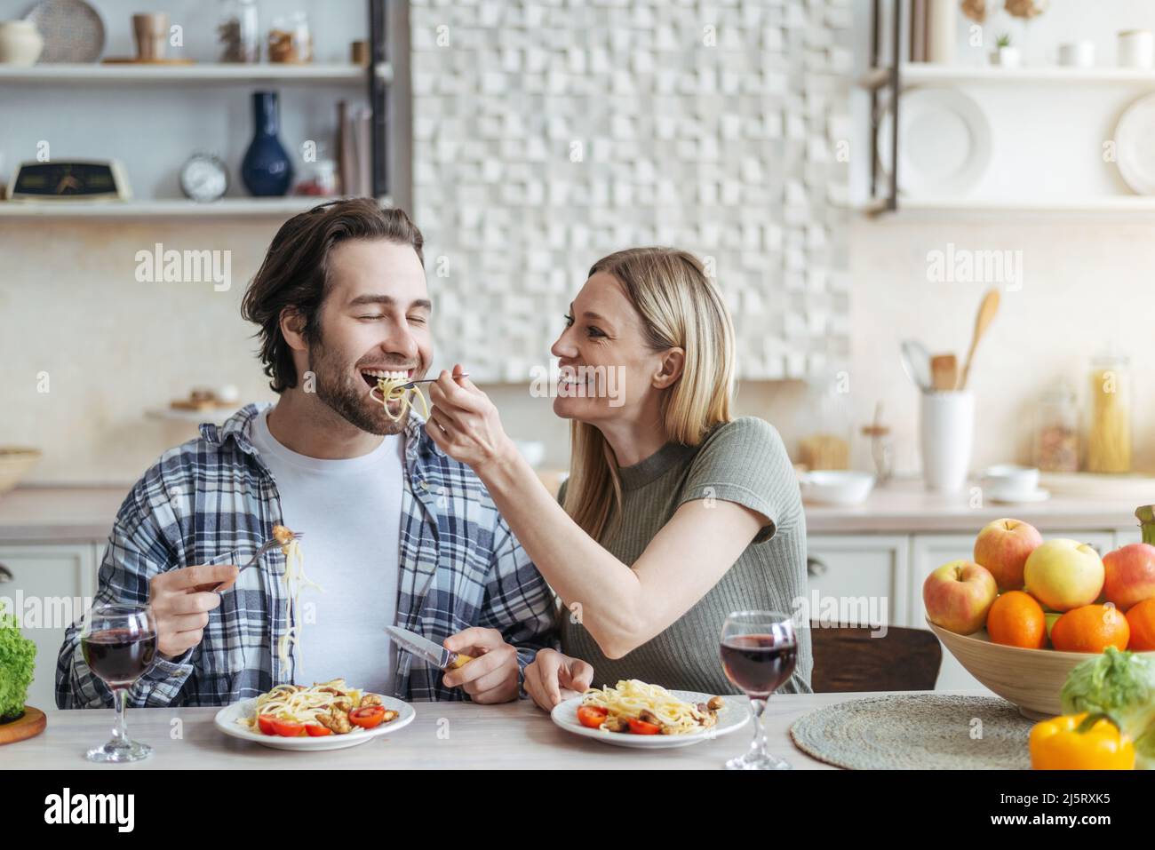 Cheerful young european woman feeds her husband of spaghetti with vegetables or pasta in minimalist kitchen Stock Photo