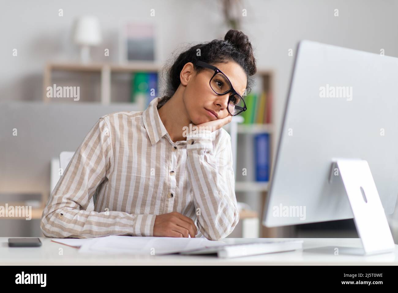 Portrait of bored businesswoman sitting at desk, leaning head on hand and looking at computer monitor Stock Photo