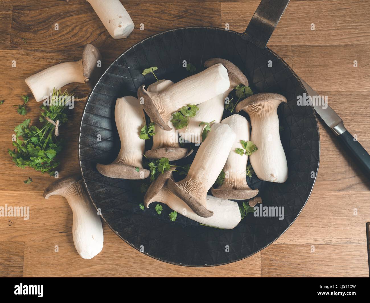 king oyster mushrooms with fresh herbs in black iron pan on a wooden cutting kitchen board decorated with parsely Stock Photo