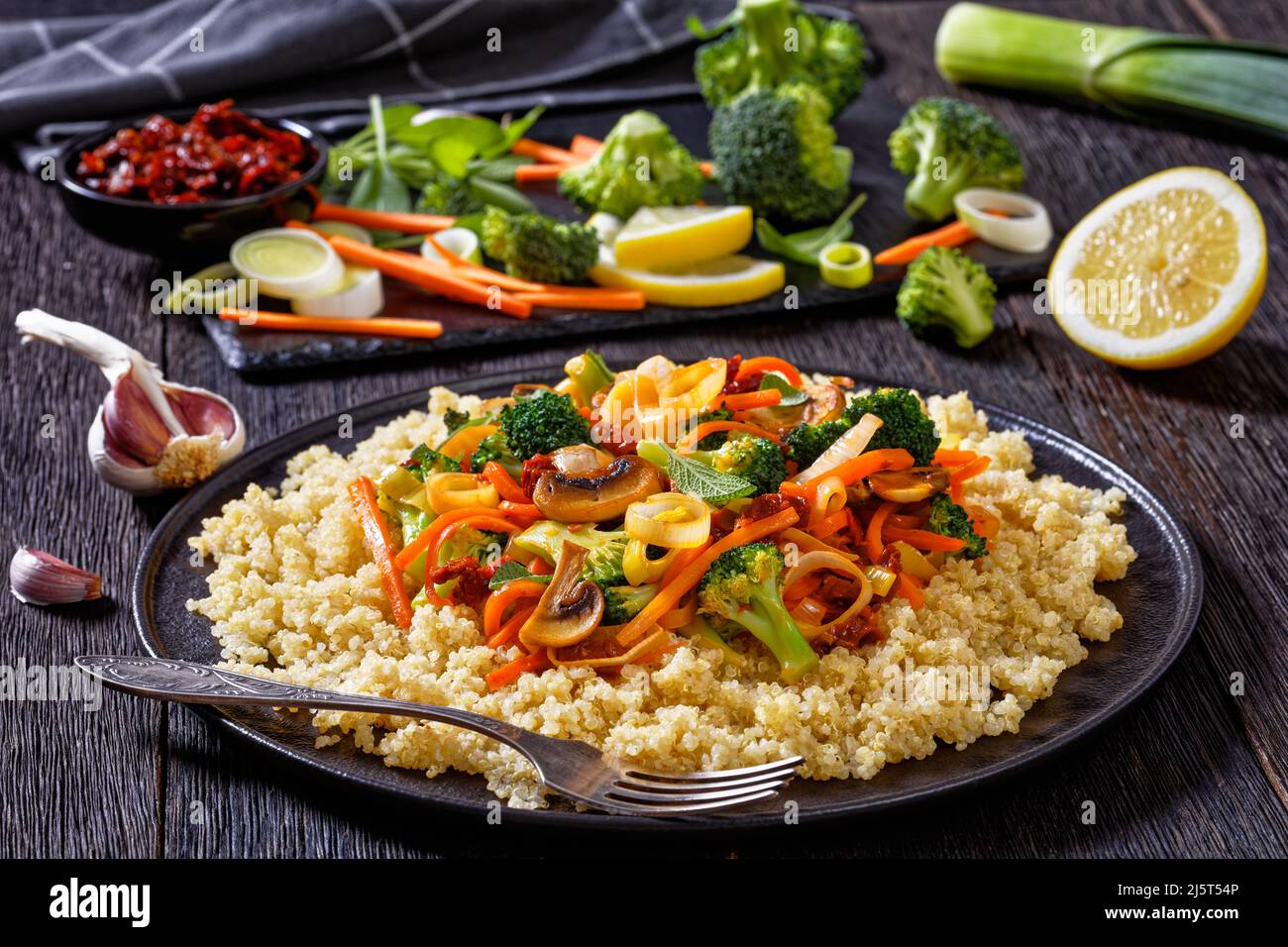quinoa topped with stir fried broccoli, julienne carrots, sun dried tomatoes, leek and mushrooms on black plate on dark wooden table with ingredients Stock Photo