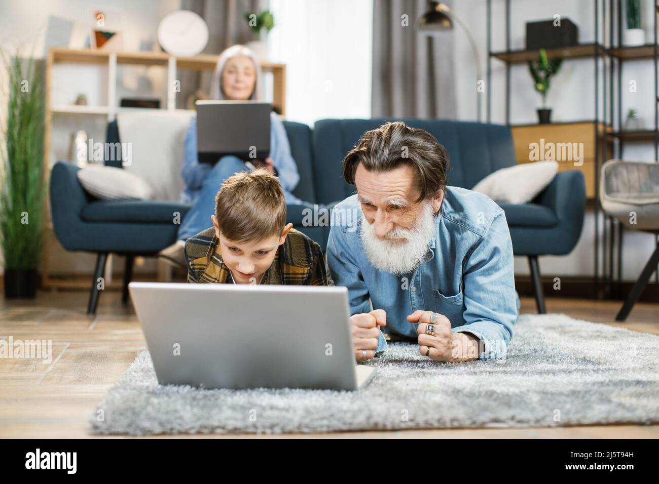 Happy different ages family leisure at home concept, mature grandmother relaxing using laptop on sofa in comfort living room while her grandchild playing on laptop with grandpa on warm floor. Stock Photo