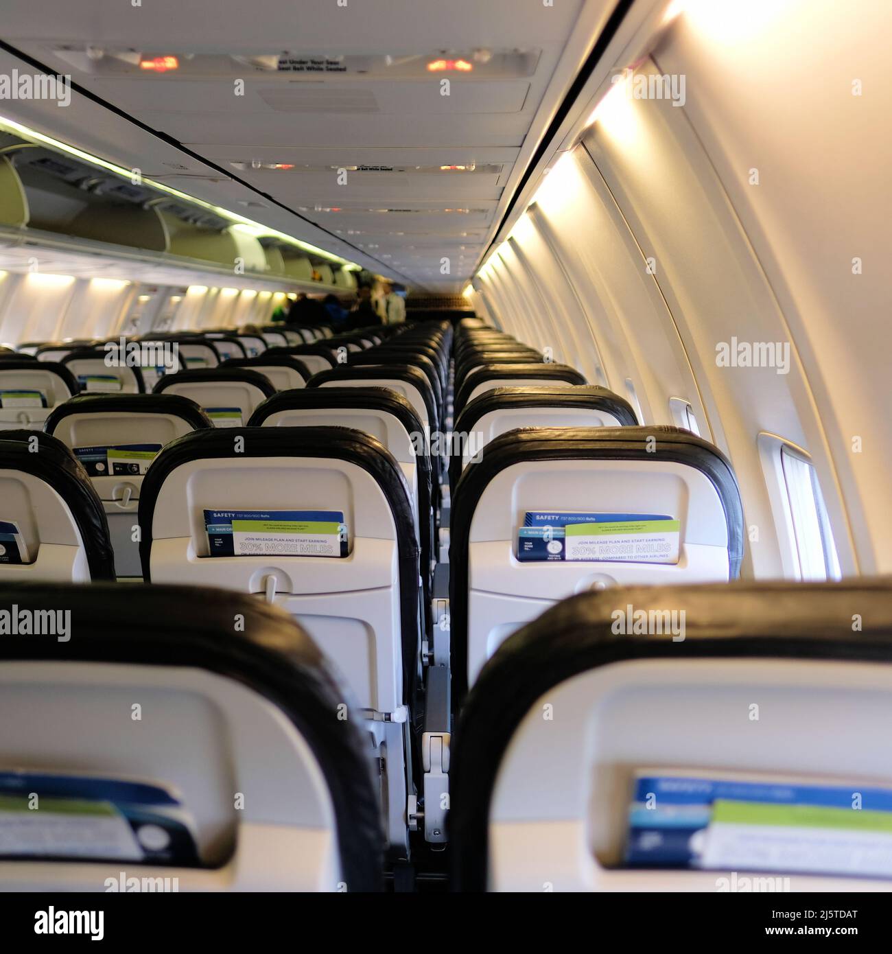 Interior cabin of an Alaska Airlines Boeing 737 with backrests, lighting panel, and passengers blurred in the distance towards the front of the plane. Stock Photo