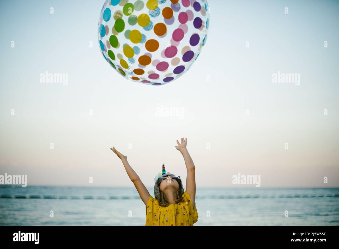 Blond seven year old girl throws rainbow ball by the ocean Stock Photo