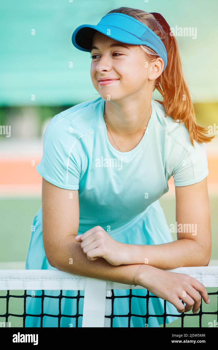 Happy portrait of a smiling teenage girl leaning on a net in a light blue sportswear. Looking to the side, posing. Lit with evening sunlight. Stock Photo