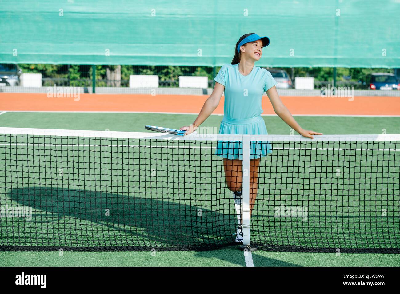 Smiley girl leaning on a tennis court net in a light blue sportswear. Looking to the side, posing. Lit with evening sunlight. Stock Photo