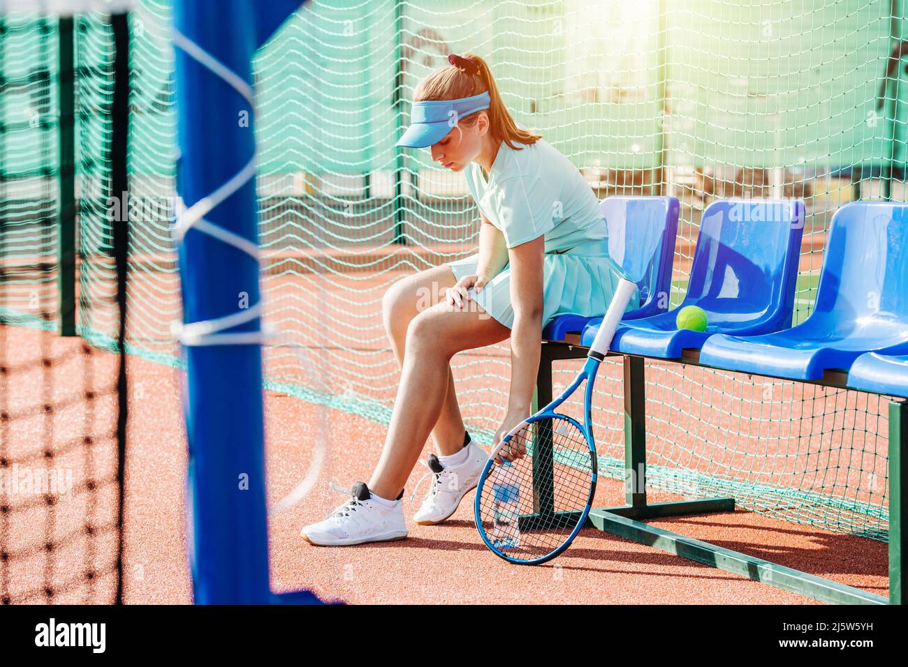 Tired weak girl resting on a plastic chair bench on an outdoor tennis court. Lit by an evening sun. High net behind her. Stock Photo