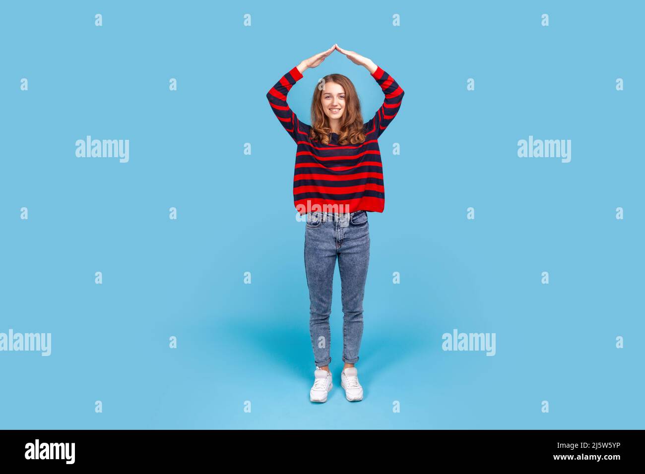Full length portrait of smiling friendly woman, doing house roof gesture over head, feeling herself in safety, wearing striped casual style sweater. Indoor studio shot isolated on blue background. Stock Photo
