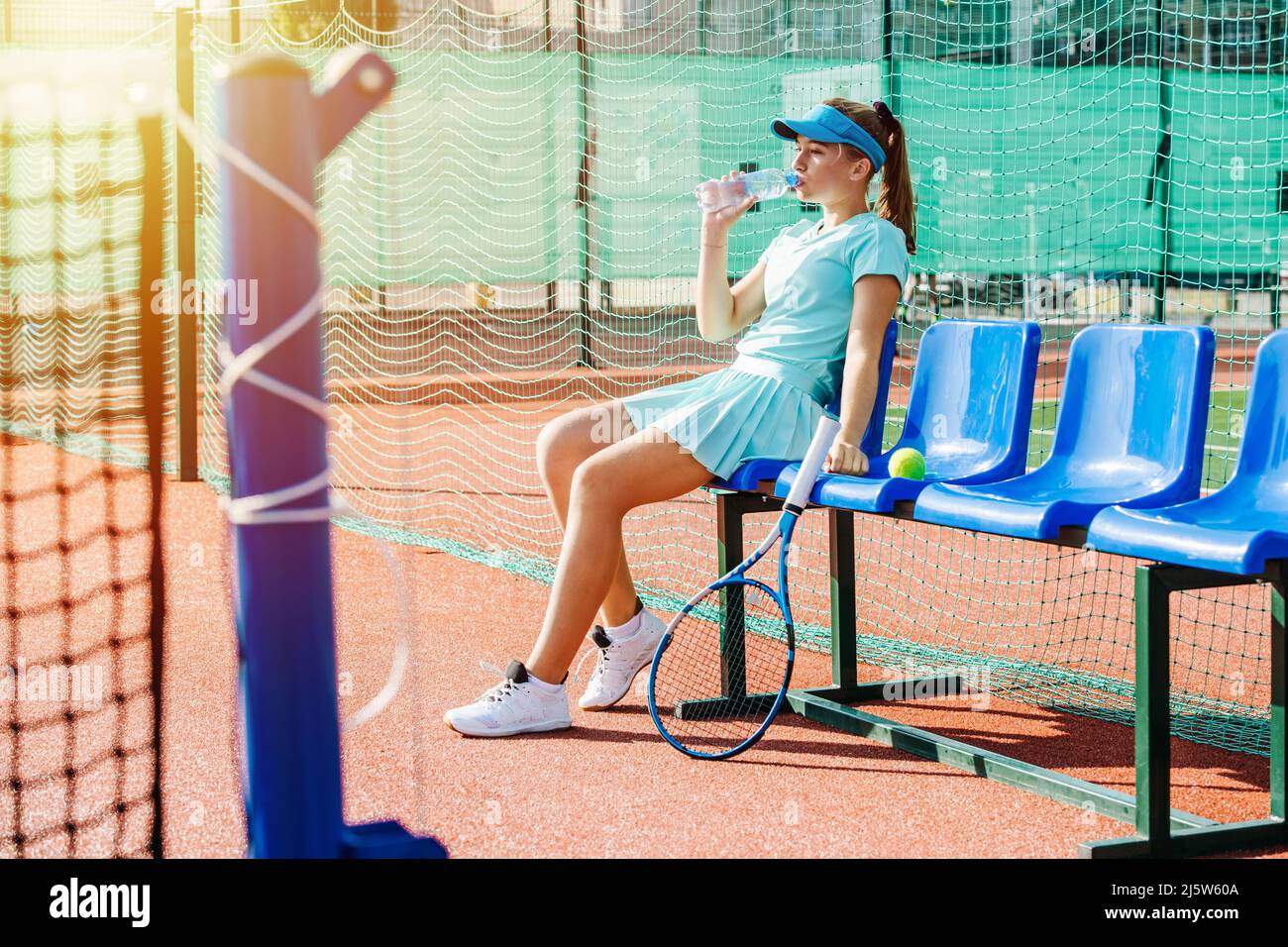 Exhausted thirsty girl resting on a plastic chair bench on an outdoor tennis court. Lit by an evening sun. High net behind her. Stock Photo