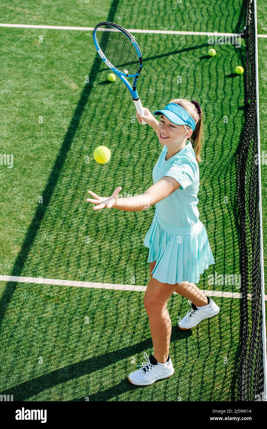 Cheerful girl practicing serve on a brand new outdoor tennis court. High angle. She is softly smiling, having a good time. Lit by an evening sun. Stock Photo