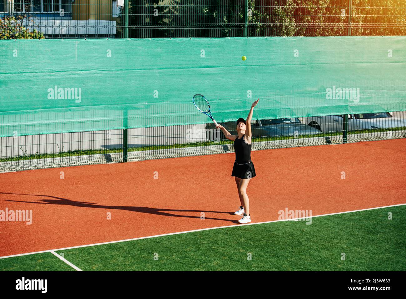 Long shot of a teenage girl practicing serve on a brand new outdoor tennis court. Vibrant green and brown with clear white lines. Stock Photo