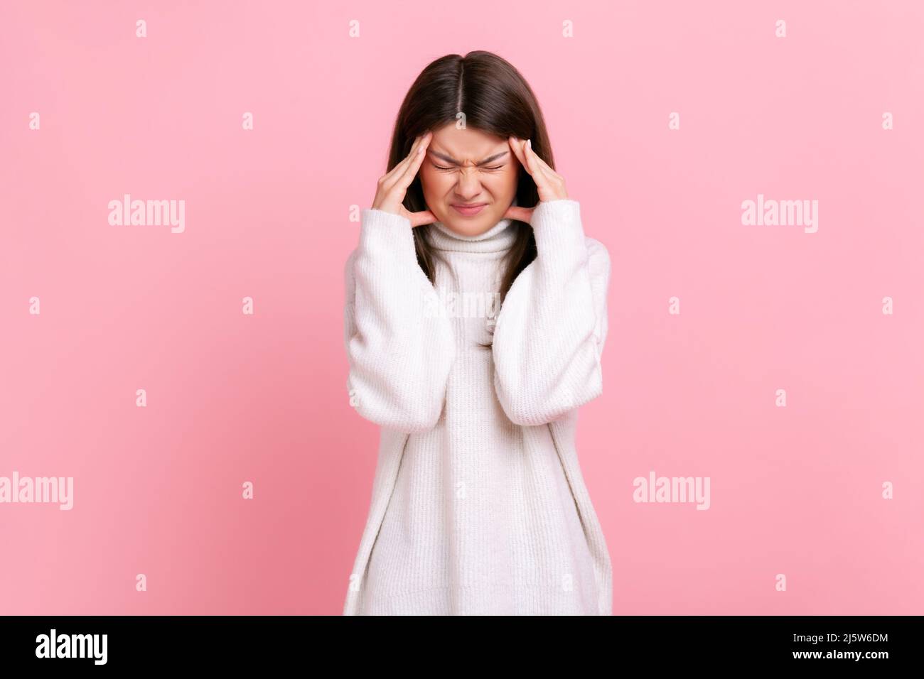 Woman frowning and clasping sore head, suffering intense headache, having migraine, fever symptoms, wearing white casual style sweater. Indoor studio shot isolated on pink background. Stock Photo