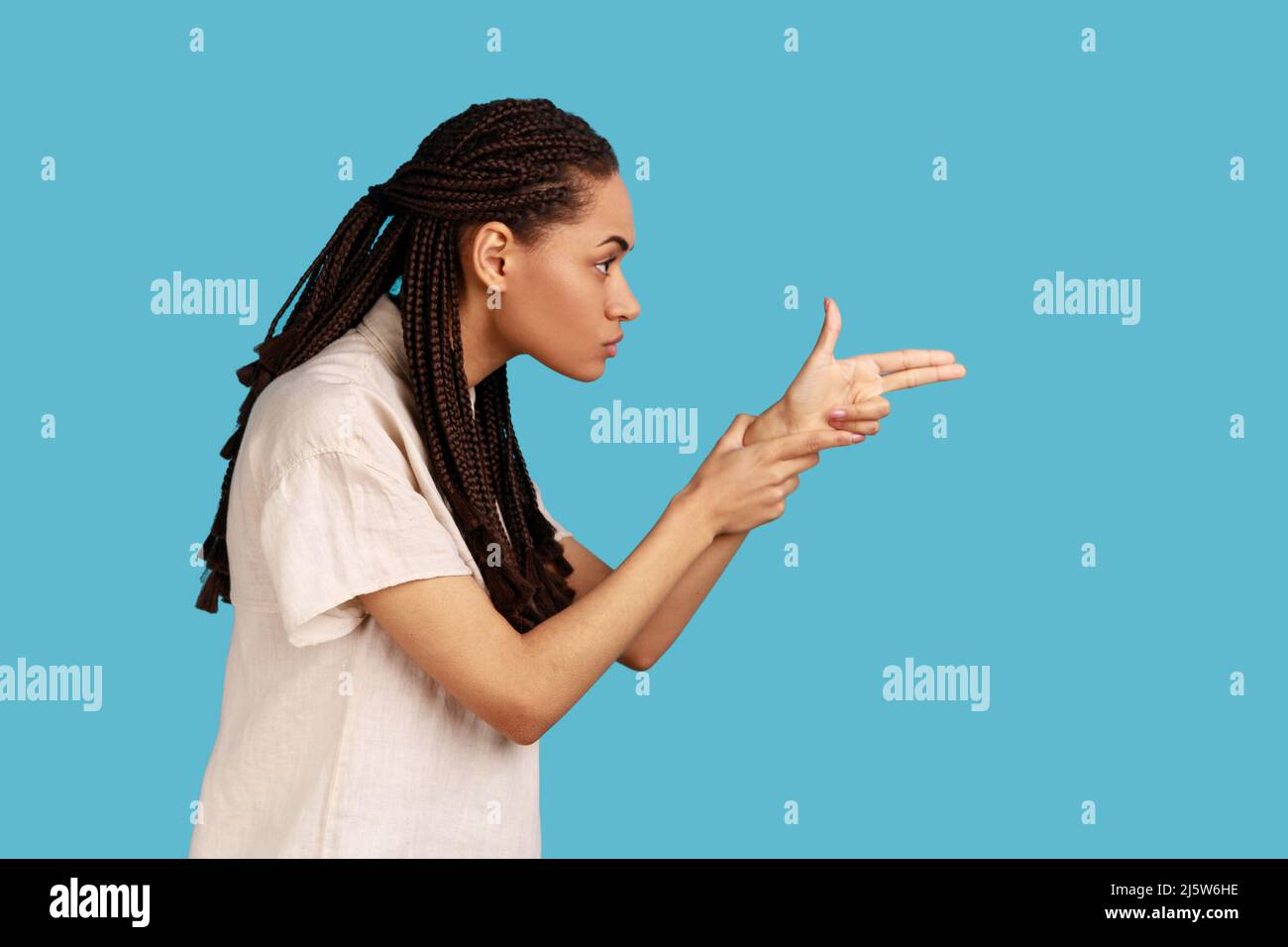 Side view of woman with black dreadlocks pointing finger gun, aiming and threatening to shoot with pistol hand gesture, wearing white shirt. Indoor studio shot isolated on blue background. Stock Photo
