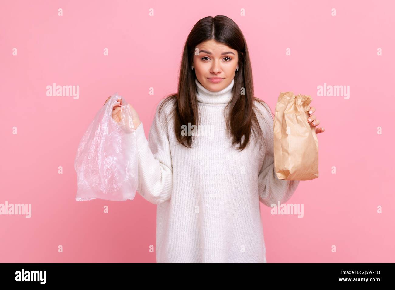 Brunette young woman holding in hands paper and poly bag, needs to make right choice to save planet, wearing white casual style sweater. Indoor studio shot isolated on pink background. Stock Photo