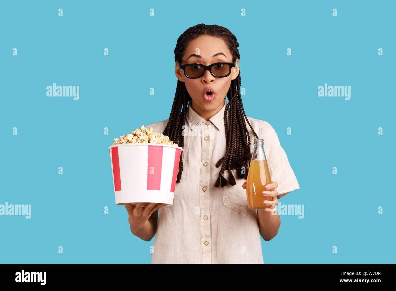 Extremely astonished woman with dreadlocks in 3d imax glasses watching movie film, hold popcorn and drink, watching amazing movie, wearing white shirt. Indoor studio shot isolated on blue background. Stock Photo