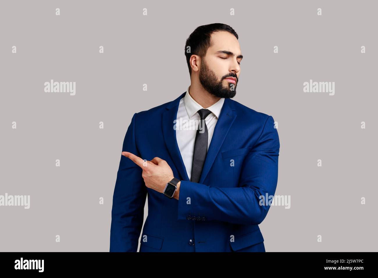 Portrait of bearded man pointing to the side and turning away with angry vexed face, giving order to leave, wearing official style suit. Indoor studio shot isolated on gray background. Stock Photo