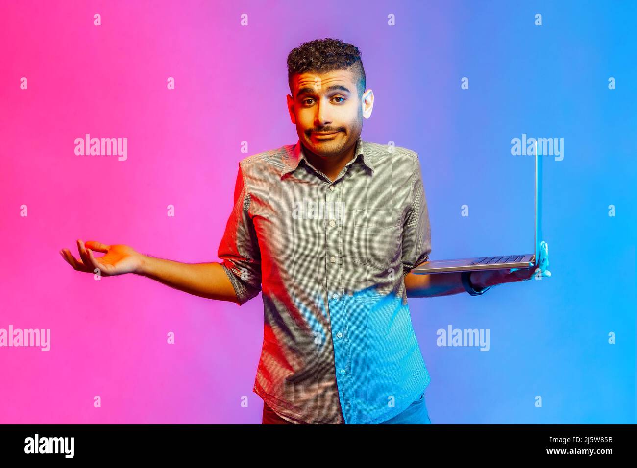 Portrait of puzzled handsome man in shirt standing holding laptop and shrugging, don't know what to do with new project. Indoor studio shot isolated on colorful neon light background. Stock Photo