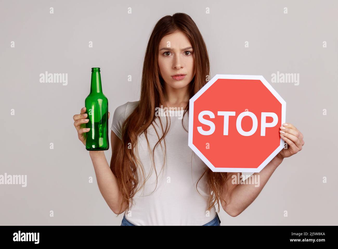 Portrait of anxious beautiful woman showing alcoholic beverage beer bottle and stop sign, warning and worrying, wearing white T-shirt. Indoor studio shot isolated on gray background. Stock Photo