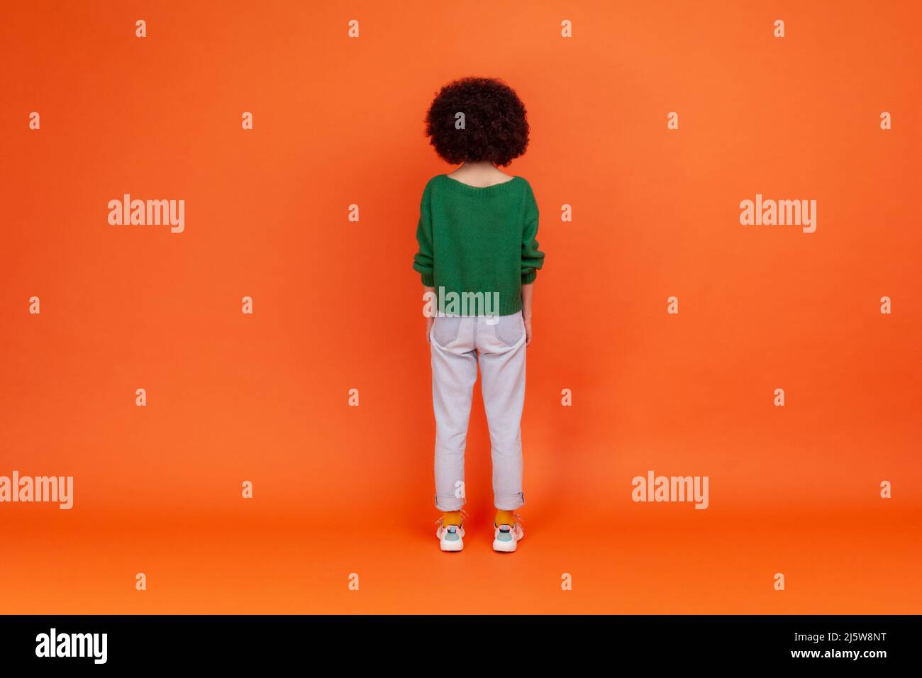 Back view of slim woman with Afro hairstyle wearing green casual style sweater and jeans standing and looking at something, waiting. Indoor studio shot isolated on orange background. Stock Photo