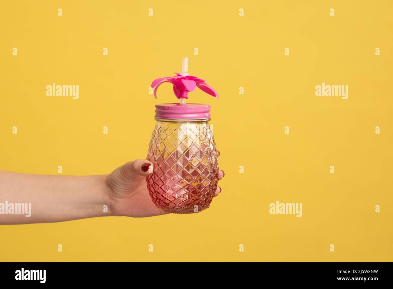 Closeup side view shot of woman hand holding pink glass jar mug with straw for cocktail or smoothie. Indoor studio shot isolated on yellow background. Stock Photo