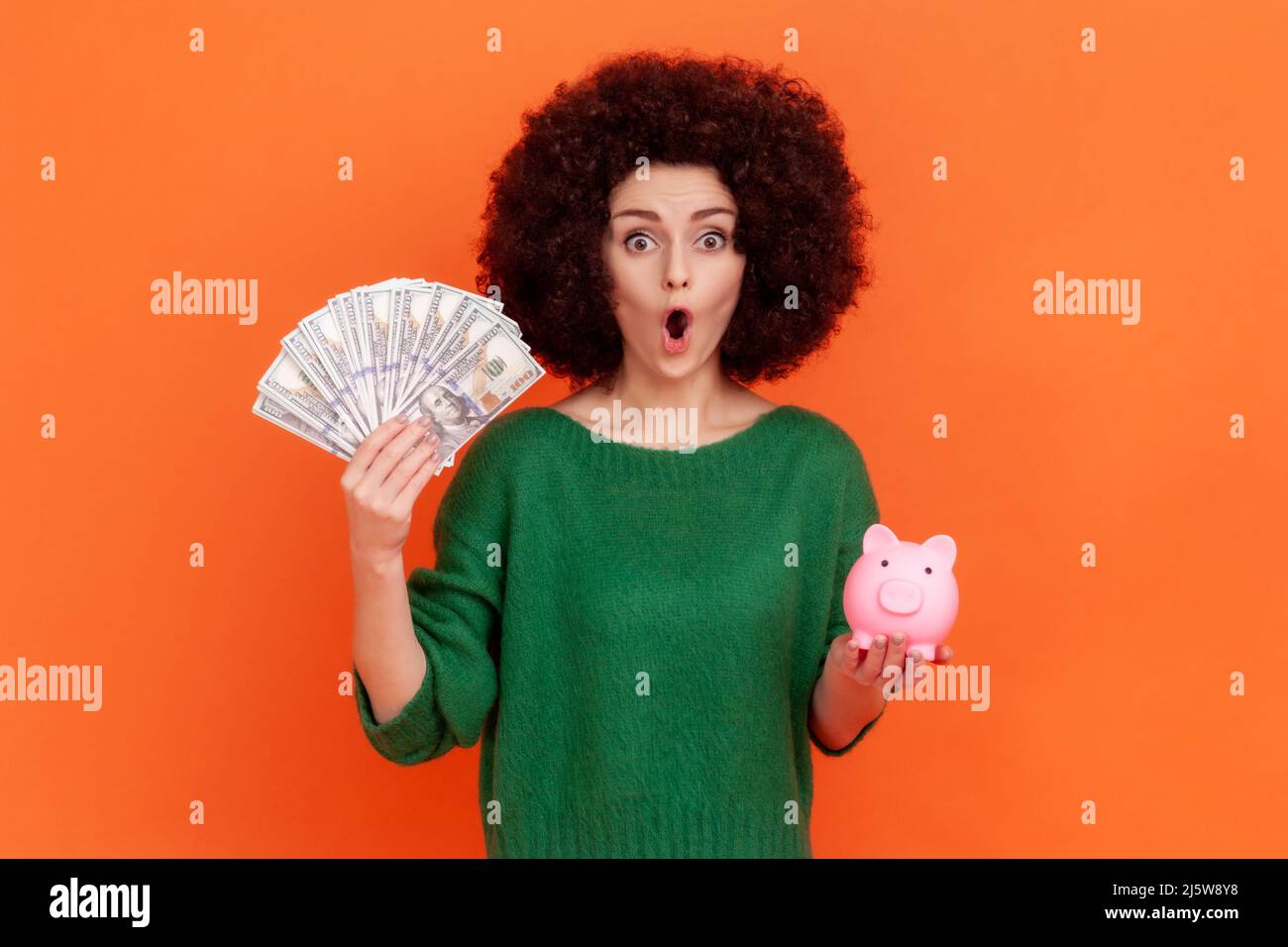 Surprised woman with Afro hairstyle in green sweater holding fan of dollars and piggy bank in hands, shocking conditions of financial investments. Indoor studio shot isolated on orange background. Stock Photo