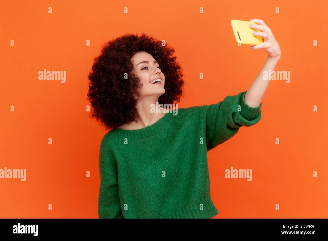 Happy friendly blogger woman with Afro hairstyle wearing green sweater broadcasting livestream, talking with followers with positive expression. Indoor studio shot isolated on orange background. Stock Photo