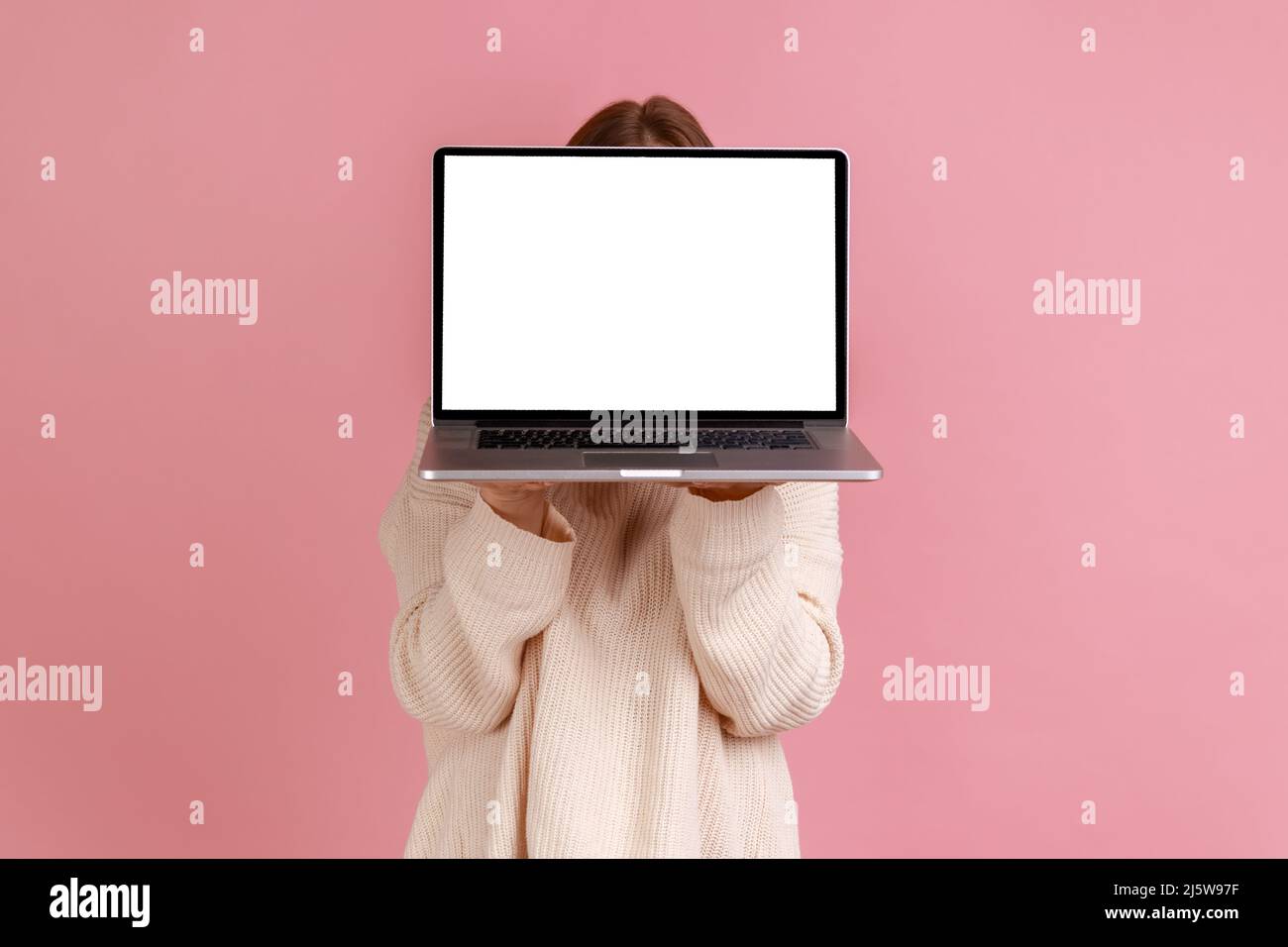 Portrait of blond woman holding laptop in hands, hiding her face behind notebook with empty display with copy space, wearing white sweater. Indoor studio shot isolated on pink background. Stock Photo