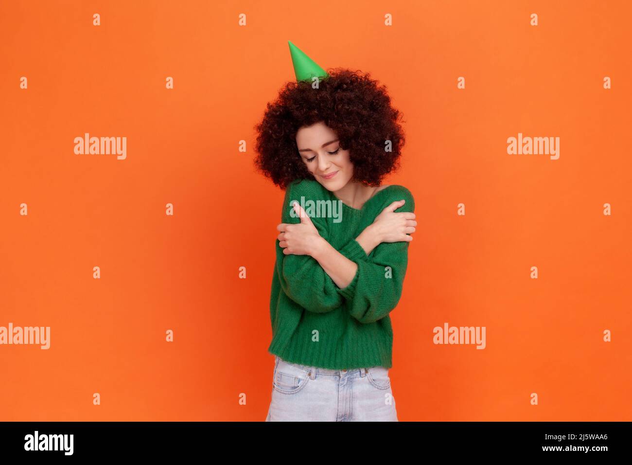 Kind birthday woman with Afro hairstyle wearing green casual style sweater and party cone, celebrating holiday, hugging herself, egoist. Indoor studio shot isolated on orange background. Stock Photo