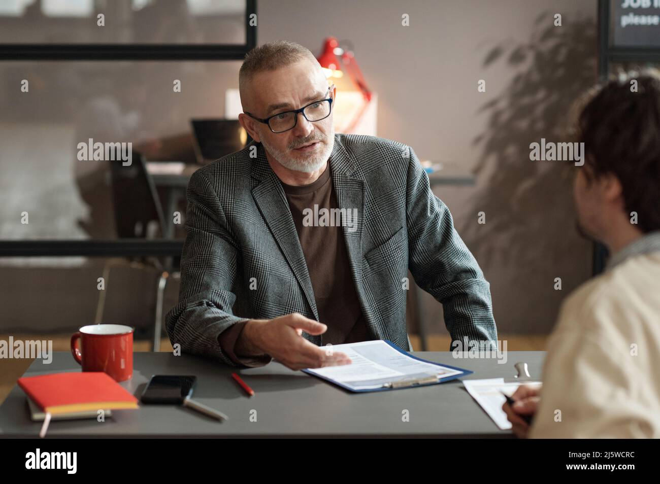 Mature businessman in eyeglasses discussing work experience in resume with candidate during job