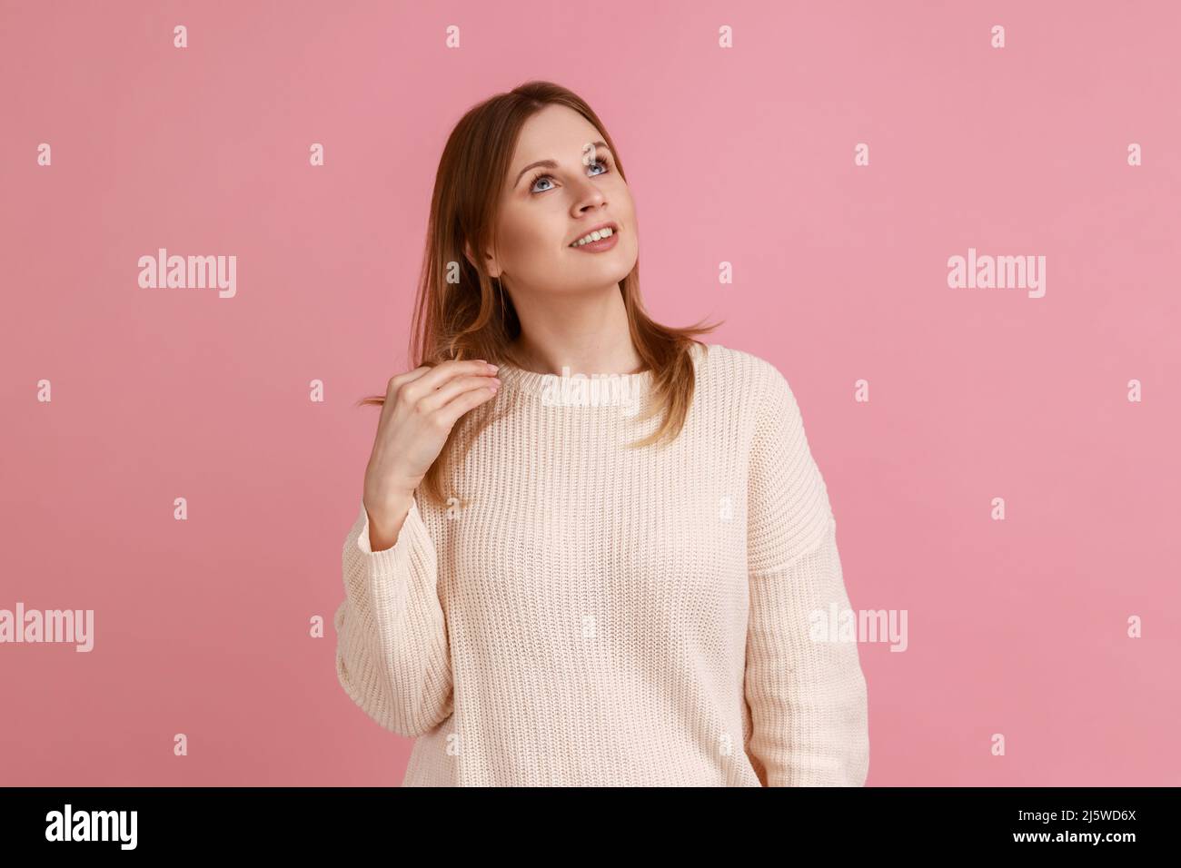 Portrait of blond woman touching hair while thinking, dreaming with joyous smiling face, pleasant fantasy, positive hopes, wearing white sweater. Indoor studio shot isolated on pink background. Stock Photo