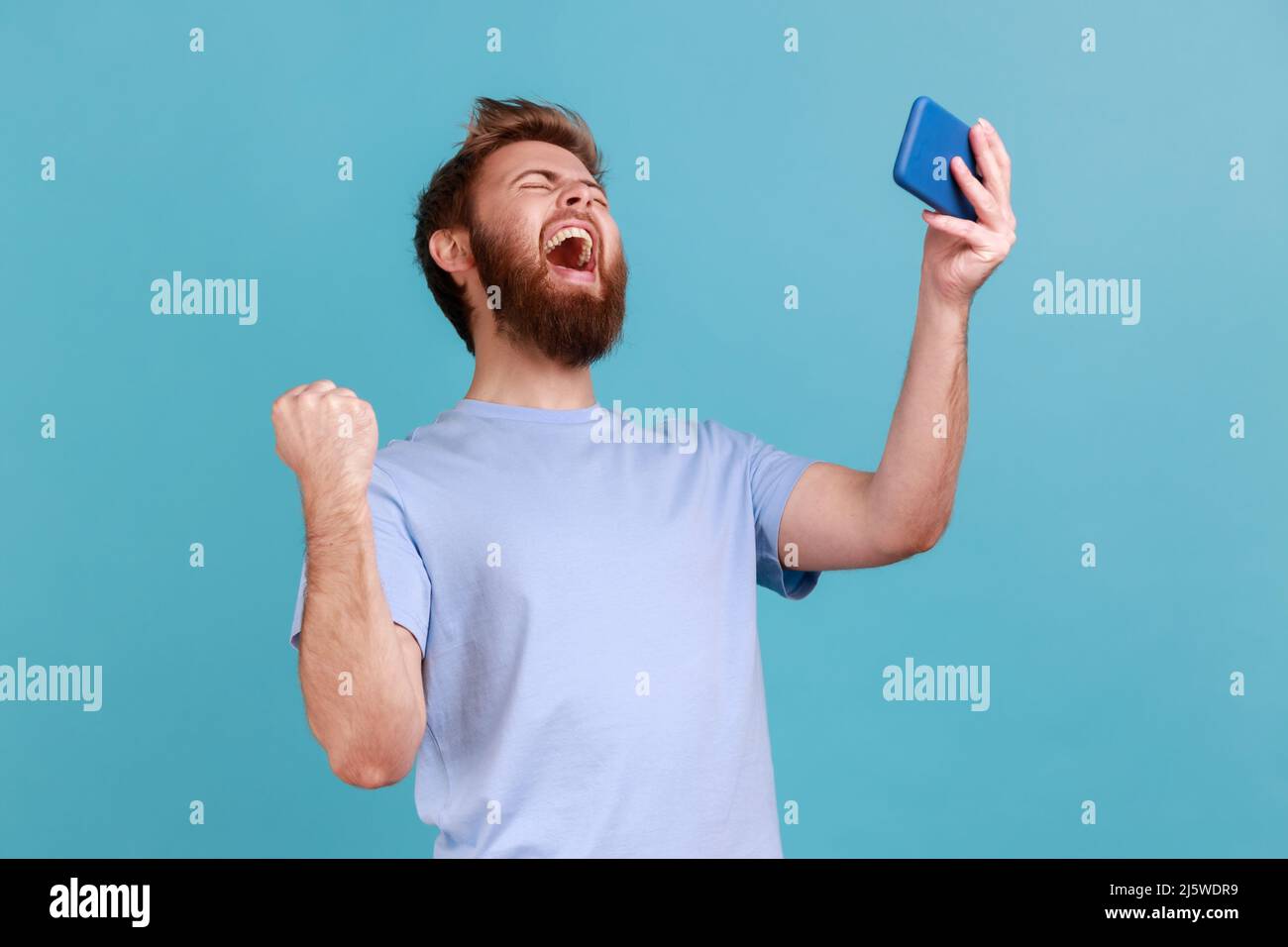 Portrait of extremely happy handsome bearded man gamer playing video game on mobile phone, celebrating victory, complete level. Indoor studio shot isolated on blue background. Stock Photo