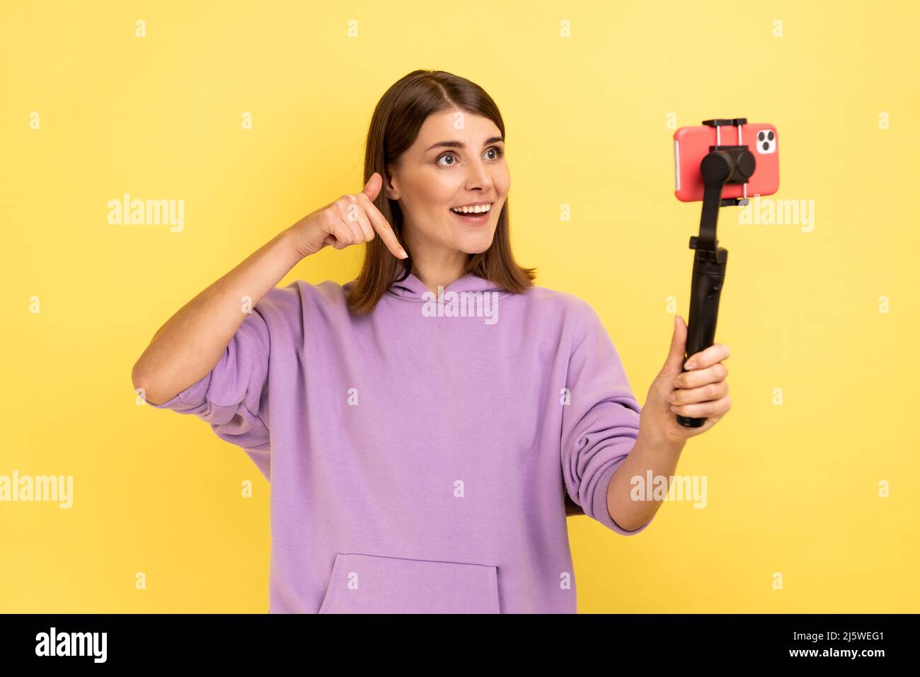 Portrait of woman using cell phone and steadicam for broadcasting livestream, pointing down, asking to subscribe, wearing purple hoodie. Indoor studio shot isolated on yellow background. Stock Photo