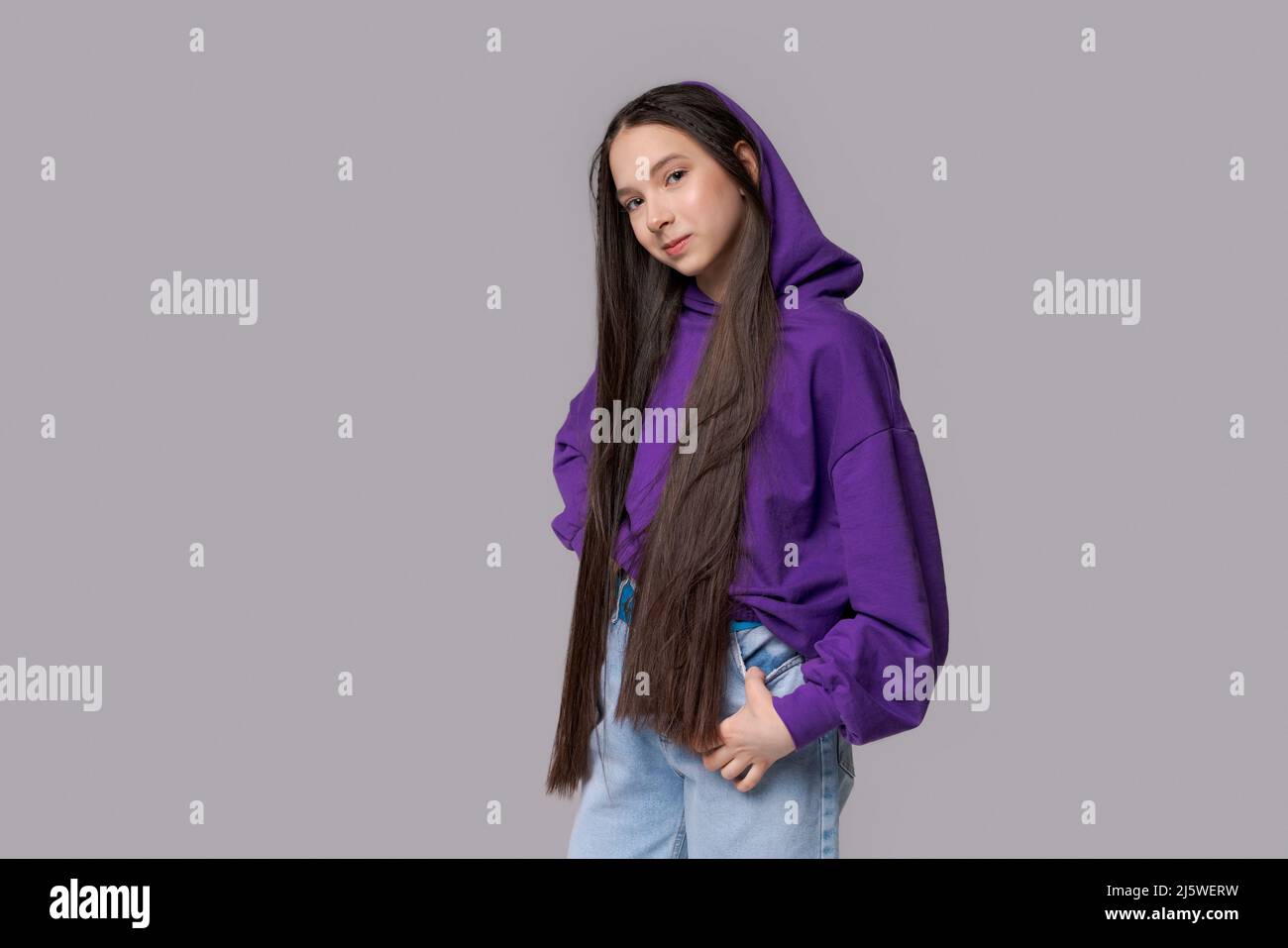 Nice cute young girl with long hair in bright hood posing on gray background. Charming girl in a lilac sweatshirt smiles and poses on isolated. Stock Photo