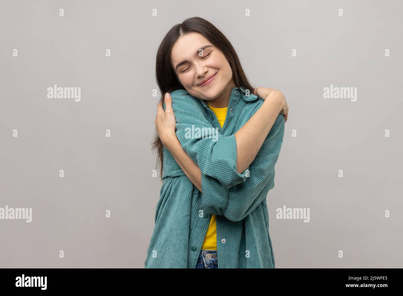 Positive self-esteem. Woman embracing herself and smiling with expression of pleasure, being selfish and narcissistic, wearing casual style jacket. Indoor studio shot isolated on gray background. Stock Photo