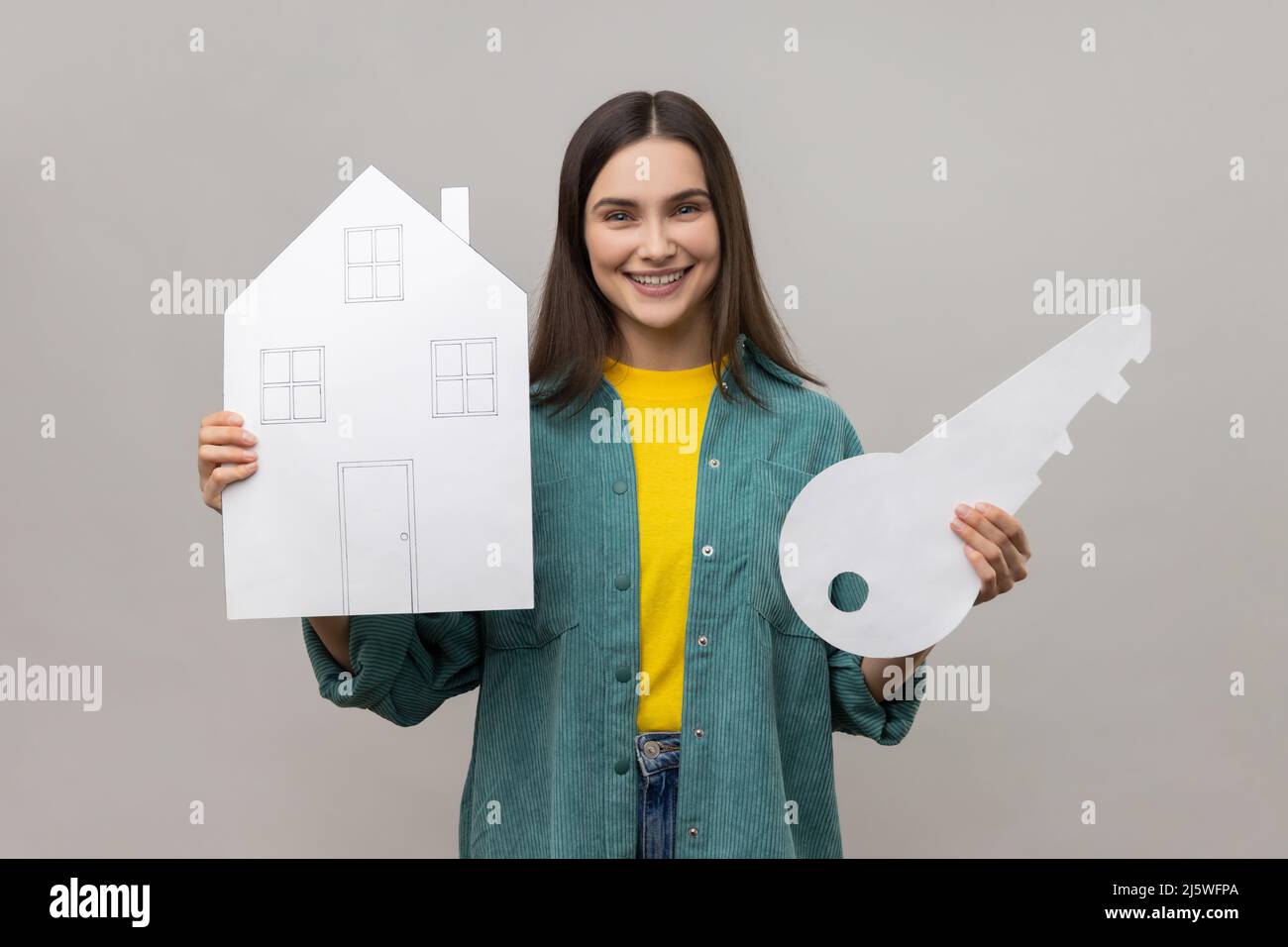 Portrait of smiling woman holding big key and paper house, looking at camera with smile, purchase of real estate, wearing casual style jacket. Indoor studio shot isolated on gray background. Stock Photo
