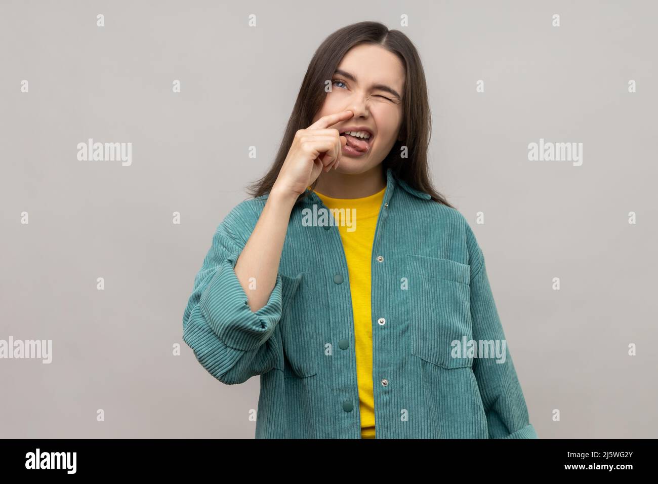 Portrait of funny woman putting finger into her nose and showing tongue, fooling around, bad habits, disrespectful behavior, wearing casual style jacket. Indoor studio shot isolated on gray background Stock Photo