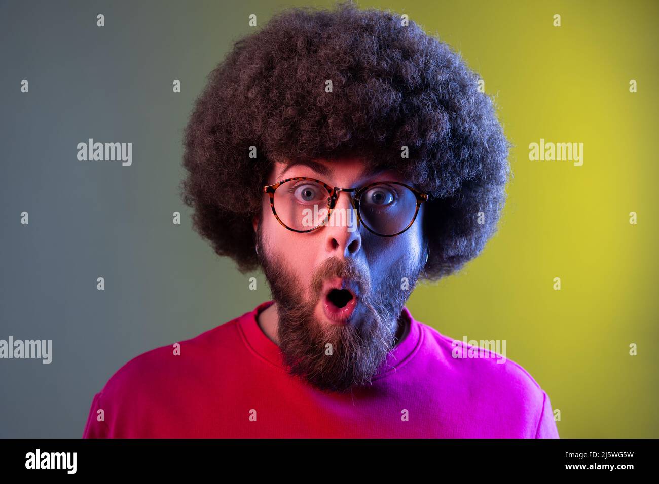Hipster man with Afro hairstyle standing with mouth open in surprise, has shocked expression, unbelievable news, wearing red sweatshirt. Indoor studio shot isolated on colorful neon light background. Stock Photo