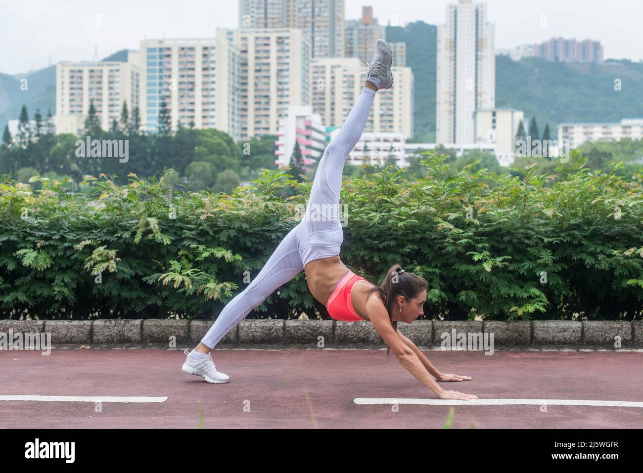 Young slim woman doing yoga down dog split exercise in city park on summer day. Female athlete stretching in standing position with arm support Stock Photo