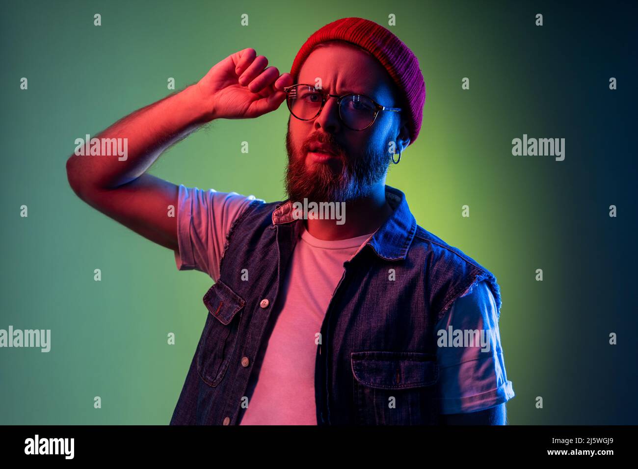 Serious hipster man looking at camera with attentive look, keeping his arms on frame of glasses, wearing beanie hat and denim vest. Indoor studio shot isolated on colorful neon light background. Stock Photo