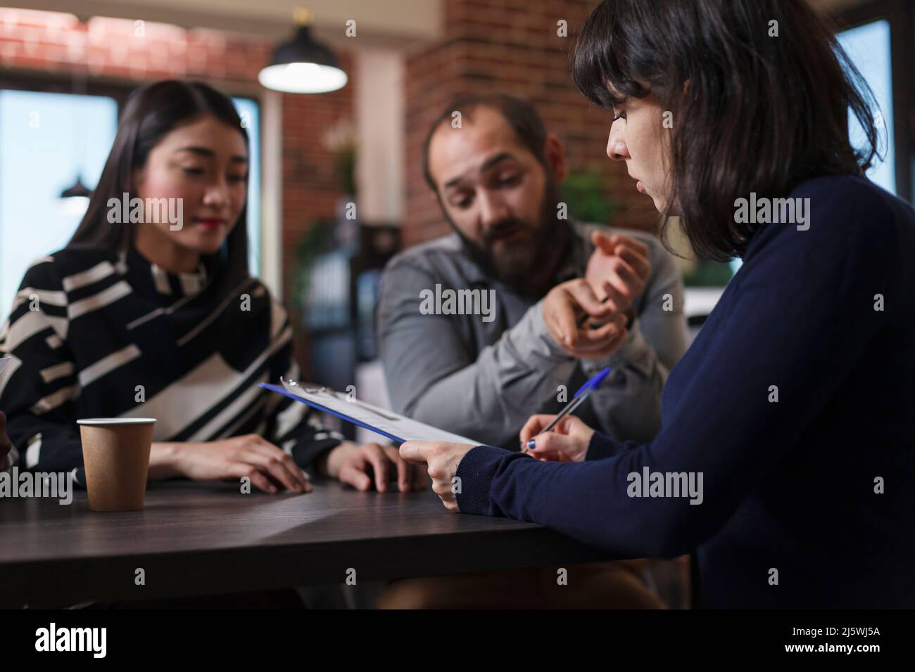 Young adult recruiter sitting at desk in office space while reviewing job applicants CVs. Multiethnic candidates talking with startup company interviewer while talking about career oportunities. Stock Photo